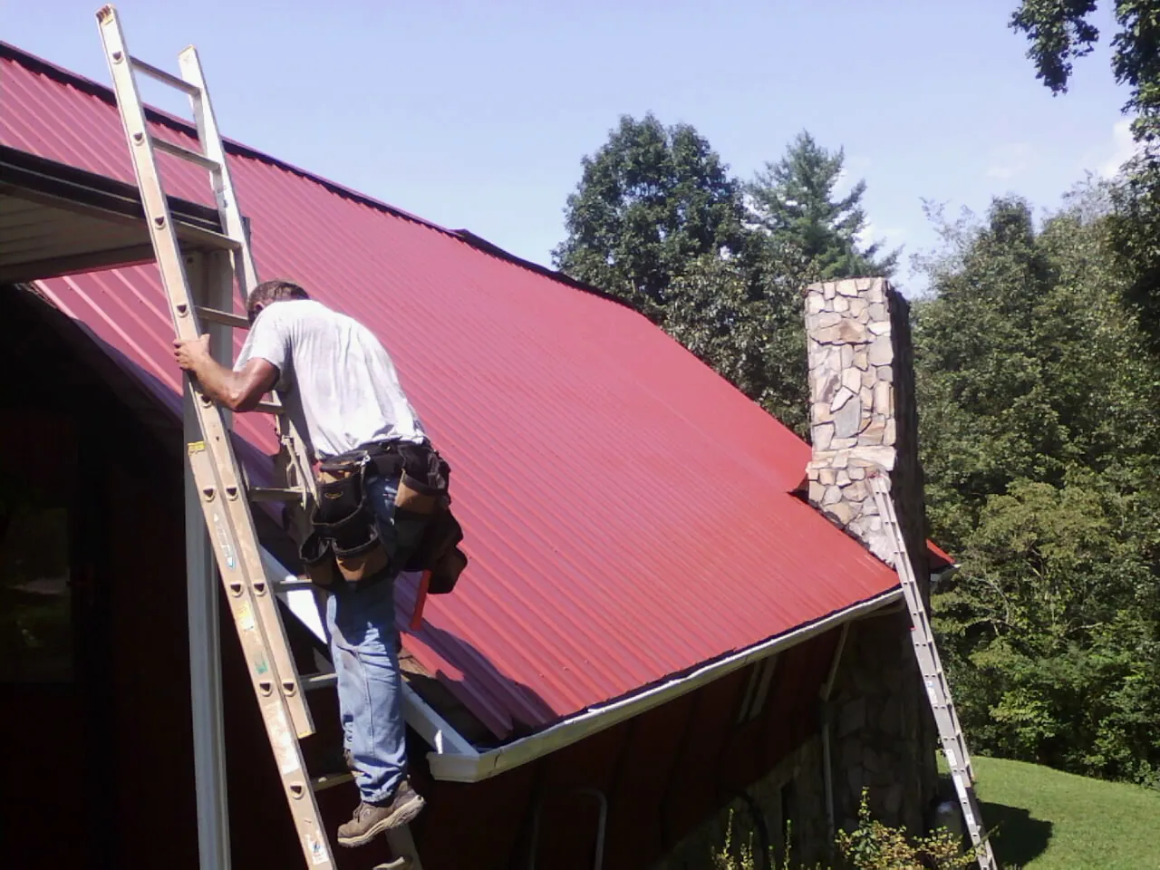 Roofer on a ladder beside a red metal roof and stone chimney — Soffit & Fascia Repair by Roofing Brothers in Fort Defiance